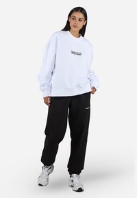Witte oversized sweatshirt met geribbelde mouwen, voorzien van zwarte geborduurde tekst op de borst. Combinatie met losse zwarte broek en sneakers.