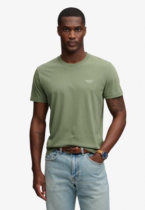 Mężczyzna w jasnozielonej koszulce Superdry, jasnoniebieskich dżinsach z brązowym plecionym paskiem i zegarkiem, stoi z jedną ręką w kieszeni.