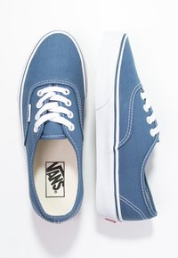 Sininen Vans-kangaslenkkari, jossa on valkoiset nauhat ja kumipohjat, yksi kenkä näkyy ylhäältä päin ja toinen sivusta päin valkoista taustaa vasten.