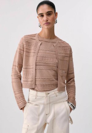 Donna con cardigan beige lavorato a maglia e top, pantaloni a vita alta color crema, orecchini a cerchio d'argento e braccialetto d'argento su sfondo neutro.