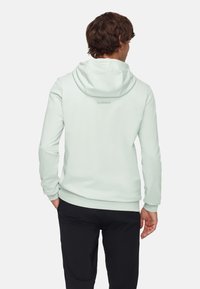 Helles mintgrünes Hoodie mit weicher Textur, langen Ärmeln, einer Kordelzugkapuze und dezentem Branding auf der Rückseite. Kombiniert mit schwarzen Hosen.