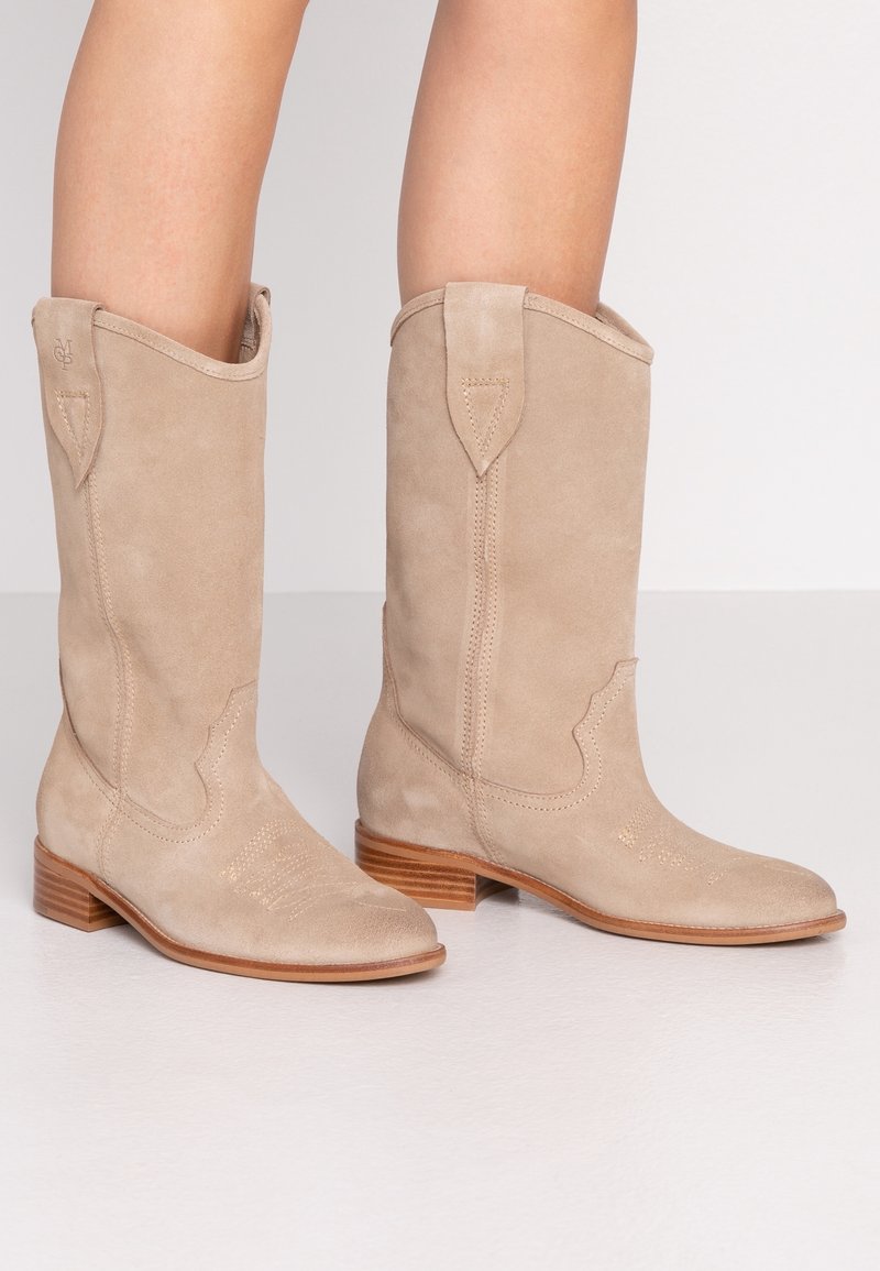 Bottes de cowboy en suede beige avec un petit talon empilé, une couture décorative, une pointe effilée et des accents de style western sur la tige.