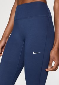 Marineblauwe leggings met hoge taille gemaakt van rekbare stof. Voorzien van een wit Nike-logo aan de linkerkant en subtiele naaddetails.