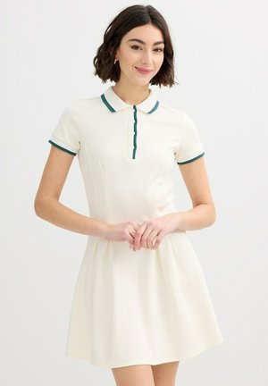 Joven mujer con cabello oscuro corto lleva un vestido crema de manga corta con cuello y puños adornados en verde, de pie frente a un fondo blanco sencillo.