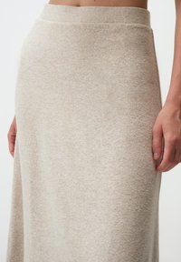 Jupe en tricot beige, arrivant au genou, avec une texture douce et un léger éclat. Elle présente une taille haute et une silhouette en A, sans coutures visibles.