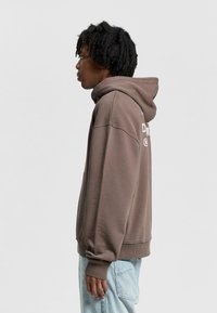 Brown hooded sweatshirt with a front pocket, cuffs, and a relaxed fit. Features graphic text on the back, made of soft, textured fabric.
