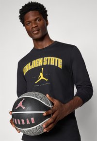 Svart langermet skjorte med "GOLDEN STATE" i gult og en gul Jumpman-logo. Holder en svart og grå teksturert basketball med en rød logo.