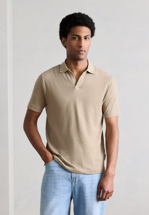 Joven de pie contra una pared lisa, vistiendo una camisa polo beige de manga corta y vaqueros azul claro, con una mano en el bolsillo y expresión neutral.