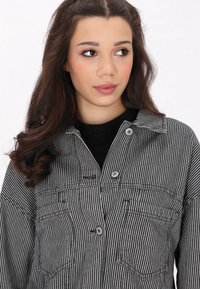 Svart och vit randig skjortjacka med krage, knappad front, två bröstfickor och en avslappnad passform. Materialet verkar lätt och strukturerat.