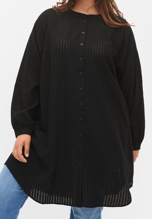 Femme portant une tunique noire à manches longues boutonnée avec une texture côtelée verticale sur un jean bleu clair, debout devant un fond uni.