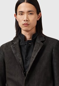 Abrigo de cuero negro con un diseño estructurado, que presenta un cuello alto, cierres de botones y una capa inferior de lana texturizada.