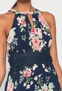 Kukalloinen mekko tummansinisellä pohjalla, jossa on vaaleanpunaisia ja vihreitä kukkia. Mekossa on avorintakuvio ja strukturoidusta tummansinisestä pitsistä valmistettu vyötärönauha.