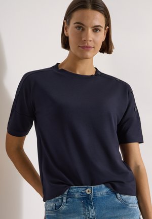Femme aux cheveux bruns courts portant un haut bleu marine à manches courtes avec des boutons sur l'épaule et un jean bleu clair.