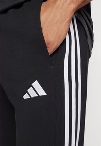 Černé sportovní kalhoty s bílým třípruhovým designem na bocích a bílým vyšitým logem; texturovaná látka s bočními kapsami.