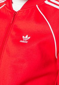 Veste zippée Adidas rouge avec des accents blancs, arborant un petit logo sur la partie gauche de la poitrine et un tissu texturé.