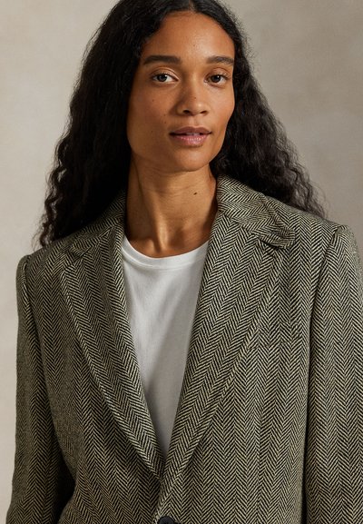 Vrouw met lang krullend haar draagt een wit shirt en een blazer met visgraatpatroon, kijkt recht in de camera met een neutrale uitdrukking.