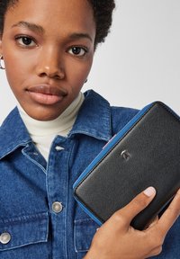 Czarny portfel o fakturowanej powierzchni z niebieskim zamkiem błyskawicznym i różowym akcentem, trzymany w ręku. W tle widoczny kołnierz dżinsowej kurtki oraz biały golf.