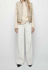 Beżowa kamizelka puchowa na jasnym swetrze, zestawiona z białymi, szerokimi spodniami z wysokim stanem oraz czarnymi butami z noskiem w szpic. Gładka tekstura i czyste linie.