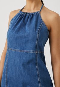 Denim halter šaty v tmavě modré barvě, s nařaseným výstřihem, bočními švy a kontrastním prošíváním. Hladká textura s přiléhavým střihem.