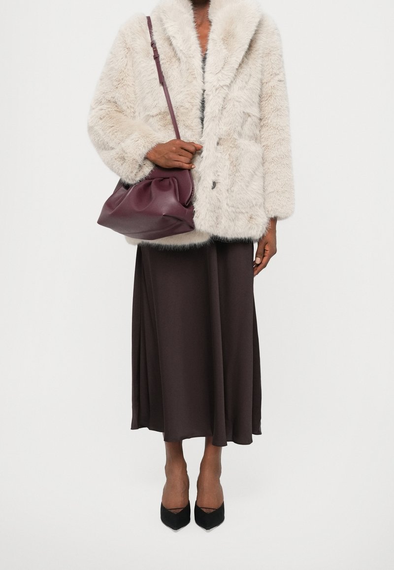 Personne portant un manteau en fausse fourrure beige clair, une longue jupe marron foncé, des talons noirs pointus et tenant un grand sac à bandoulière bordeaux.