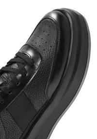 Zapatilla negra con un upper de cuero texturizado, acentos lisos, punta perforada y una suela gruesa y acolchada para mayor altura.