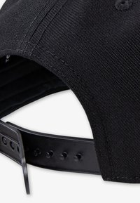 Black cap with a textured fabric, featuring a plastic adjustable strap at the back with several holes for sizing. Seam details visible.