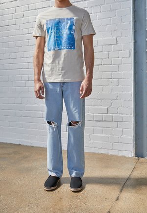 T-shirt en coton beige avec un motif graphique bleu, associé à un jean en denim clair avec des déchirures au niveau des genoux, et des chaussures noires à enfiler avec des détails en jute.