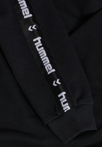 Schwarzer Sweatshirt-Stoff mit geripptem Bündchen und einem glänzenden schwarzen Seitenstreifen, der mit "hummel" in Weiß beschriftet ist und ein markantes Chevron-Muster aufweist.