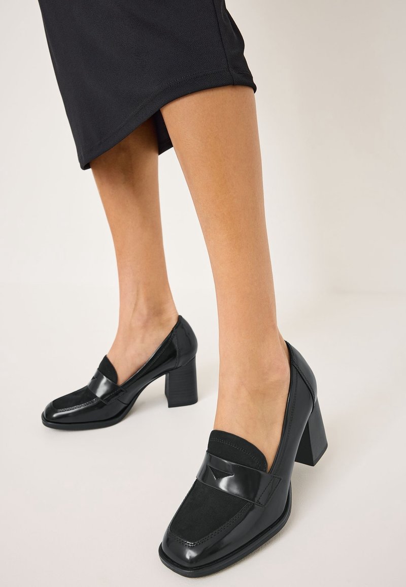Femme portant des mocassins noirs à talons carrés et une jupe noire, debout sur un sol clair devant un fond uni.