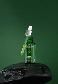 Groene glazen fles vitamine-olie met een zilveren pipetdeksel, met witte tekst "DIEPE VOCHTIGHEID [OMEGA-3]" op een zwarte stenen ondergrond.