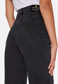 Černé džínsy s vysokým pasem vyrobené z texturované denimové tkaniny. Obsahují zadní kapsu a značkovou cedulku na pase se zlatým nápisem.