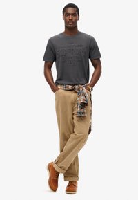 Grå T-shirt med præget logo, beige bukser og brune snørebåndsstøvler. Rutet flannel skjorte bundet omkring livet. Afslappet outfit.