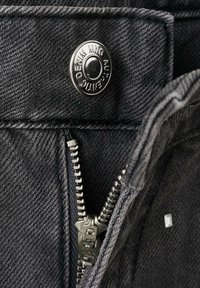 Jeans de mezclilla negra con un botón a presión plateado y detalle de cremallera YKK, con una tela texturizada y costuras visibles en la cinturilla.