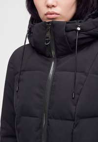 Femme portant une veste matelassée noire avec un col montant, une fermeture éclair à l’avant et des cordons sur la capuche.