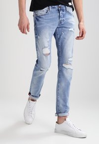 Jasnoniebieskie dżinsy z przetarciami, z podwiniętymi nogawkami i prostym krojem, w połączeniu z białymi trampkami.