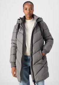 Manteau gris rembourré avec capuche, présentant des coutures diagonales, des bretelles noires et des poches zippées. Porté sur un pull à mailles torsadées crème.