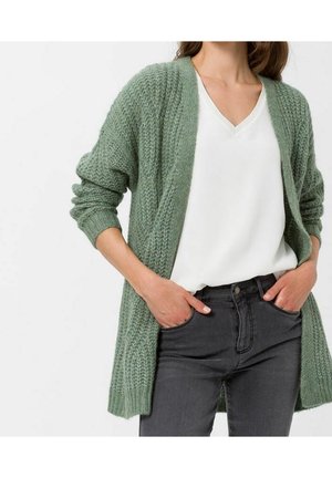 Femme portant un cardigan en maille vert par-dessus un chemisier blanc à col en V et un jean gris foncé, les mains dans les poches, debout devant un fond blanc.