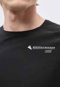 Nærbilde av en person som har på seg en svart teksturert skjorte med hvit "Klättermusen DISER"-logo på brystet.