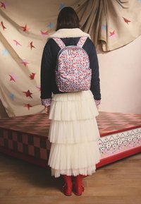 Femeie cu păr închis la culoare purtând un rucsac floral, jachetă bleumarin cu guler de blană, fustă stratificată din tul alb, colanți roșii și pantofi roșii, stând în interior.