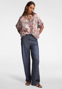 Mujer con una blusa corta de manga corta, suelta y estampada, pantalones de mezclilla oscuros de pierna ancha, sandalias marrones y joyería dorada, de pie contra un fondo blanco.