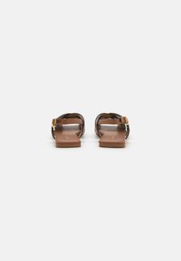 Sandale plate maro cu un design din curea împletită și detalii cu cataramă aurie. Talpa este de o culoare tan netedă, oferind un stil minimalist.