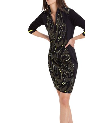 Kobieta ubrana w czarną i zieloną sukienkę o wzorze do kolan z rękawami do ćwiartki, stojąca z rękami na biodrach na białym tle.