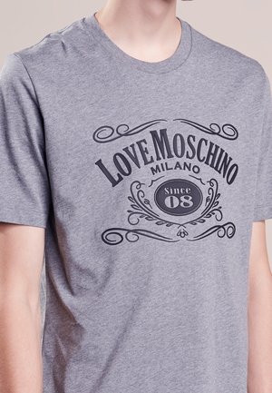 Personne portant un T-shirt gris avec un texte décoratif noir au centre de la poitrine disant « Love Moschino Milano Since 08 ».