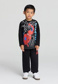 T-shirt nera a maniche lunghe con grafica di Spider-Man e dettagli rossi e blu, abbinata a pantaloni della tuta neri e scarpe sportive bianche.