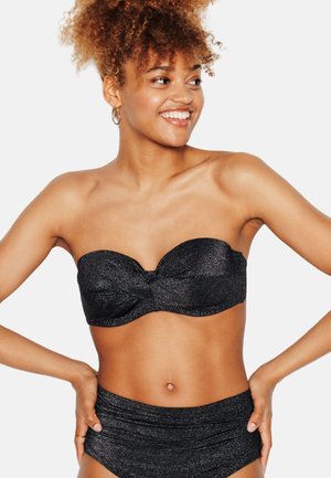 Donna sorridente con capelli ricci che indossa un top bikini bandeau nero senza spalline e slip a vita alta su uno sfondo semplice.