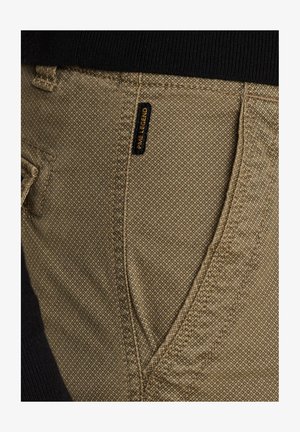 Pantalones caqui con un tejido texturizado, que cuentan con un bolsillo lateral y una etiqueta negra con letras doradas que dice "PAVE LEGEND."