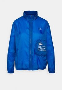 Světle modrá zipová bunda s vysokým límcem, elastickými rukávy, malým logem na hrudi a velkou kapsou na zip s bílým logem Lacoste na přední straně.