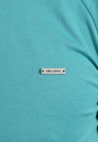 T-shirt turchese a maniche corte realizzato in tessuto morbido, con un'etichetta grigia rettangolare con la scritta "SMILODOX" cucita sul petto.