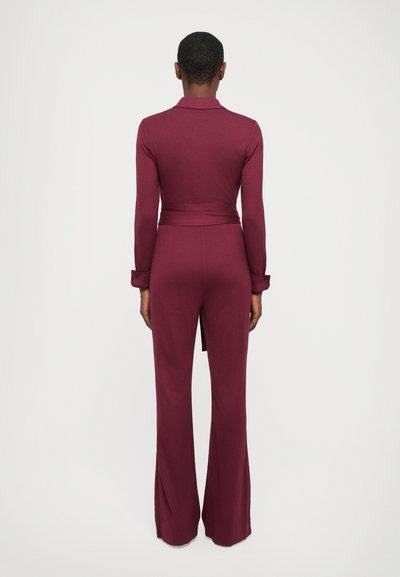 Burgundy jumpsuit with long sleeves, a fitted waist, and wide flared legs. Smooth fabric with a collar and side slits at the hem.