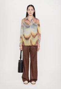 Cămașă butonată plisată, multicoloră, cu un model geometric în nuanțe pastelate. Asortată cu pantaloni largi maro și o geantă neagră.